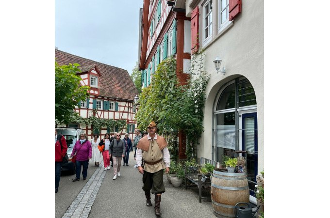 Ein Mann in historischer Kleidung geht durch eine malerische Straße in Schorndorf, flankiert von Fachwerkhäusern und einer Gruppe von Besuchern., © Eigenbetrieb Tourismus und Citymanagement Schorndorf