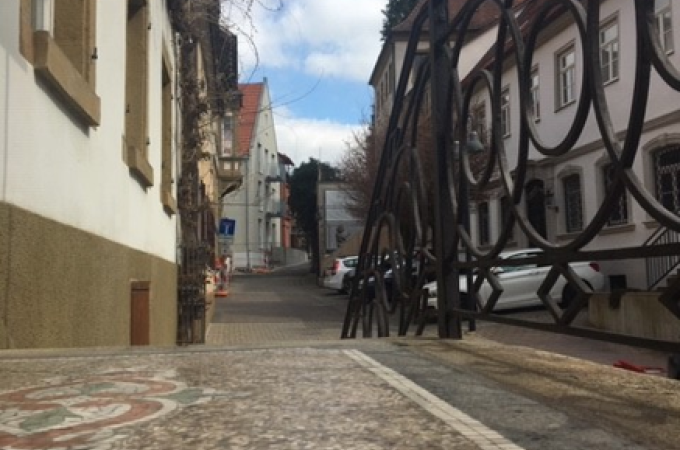 Ein kunstvolles Tor öffnet den Blick auf eine gepflasterte Straße, gesäumt von historischen Gebäuden mit roten Dächern und weißen Fassaden., © Touristik und Marketing GmbH