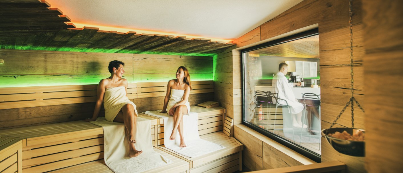 Zwei Frauen entspannen in einer modernen Sauna mit Holzverkleidung und farbiger Beleuchtung. Durch ein Fenster ist ein Mann in einem Bademantel zu sehen., &copy; &copy;LOCHER Fotodesign & Manufaktur &copy;Staatsbad Wildbad GmbH