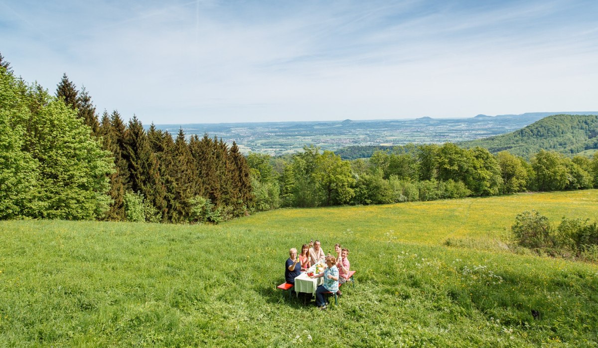 Eine Gruppe von Menschen sitzt an einem Tisch auf einer grünen Wiese. Im Hintergrund sind die Drei Kaiserberge und eine weite Landschaft zu sehen., © Landkreis Göppingen