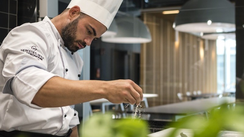 Ein Koch in weißer Uniform und Mütze lächelt in einer modernen Küche. Er hält einen großen Löffel. Oben links ist das Logo von Mercedes-Benz Gastronomie zu sehen., © Mercedes-Benz AG Ein Koch in weißer Uniform und Mütze lächelt in einer modernen Küche. Er hält einen großen Löffel. Oben links ist das Logo von Mercedes-Benz Gastronomie zu sehen., © Mercedes-Benz AG