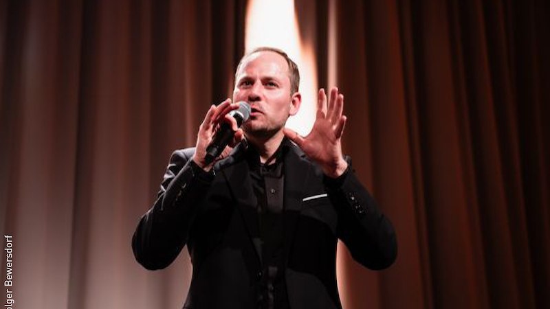 Ein Mann in schwarzem Anzug spricht leidenschaftlich in ein Mikrofon auf einer Bühne mit braunem Vorhang., © Stadt Kornwestheim Kulturmanagement Ein Mann in schwarzem Anzug spricht leidenschaftlich in ein Mikrofon auf einer Bühne mit braunem Vorhang., © Stadt Kornwestheim Kulturmanagement
