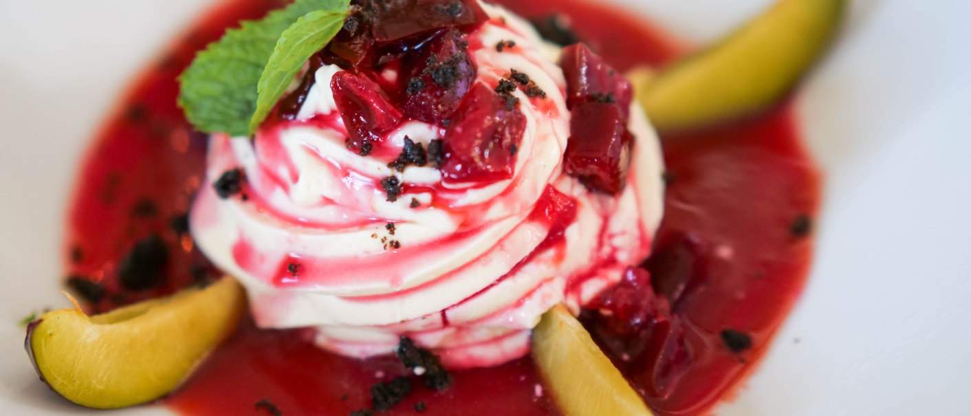 Ein kunstvoll angerichtetes Dessert mit Sahne, roten Fruchtst&uuml;cken und So&szlig;e, garniert mit Minzblatt und Pflaumenscheiben., &copy; RestaurantPflum