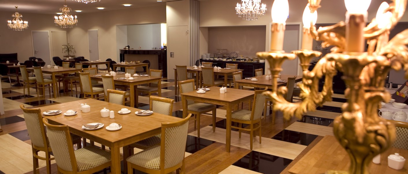 Eleganter Frühstücksraum mit Holztischen und Stühlen, dekoriert mit Kronleuchtern. Der Raum wirkt einladend und stilvoll., © Ross Messehotel Eleganter Frühstücksraum mit Holztischen und Stühlen, dekoriert mit Kronleuchtern. Der Raum wirkt einladend und stilvoll., © Ross Messehotel