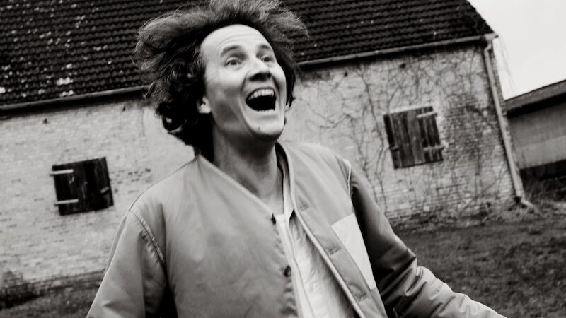 Eine Person lacht ausgelassen vor einem alten Gebäude mit Ziegeln und Fensterläden. Schwarz-weiß Fotografie., © Kulturhaus Schwanen