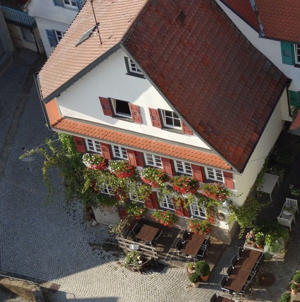 Traditionelles Geb&auml;ude mit roten Fensterl&auml;den und blumengeschm&uuml;ckter Fassade. Im gepflasterten Hof stehen Tische und St&uuml;hle bereit., &copy; cWeinstube