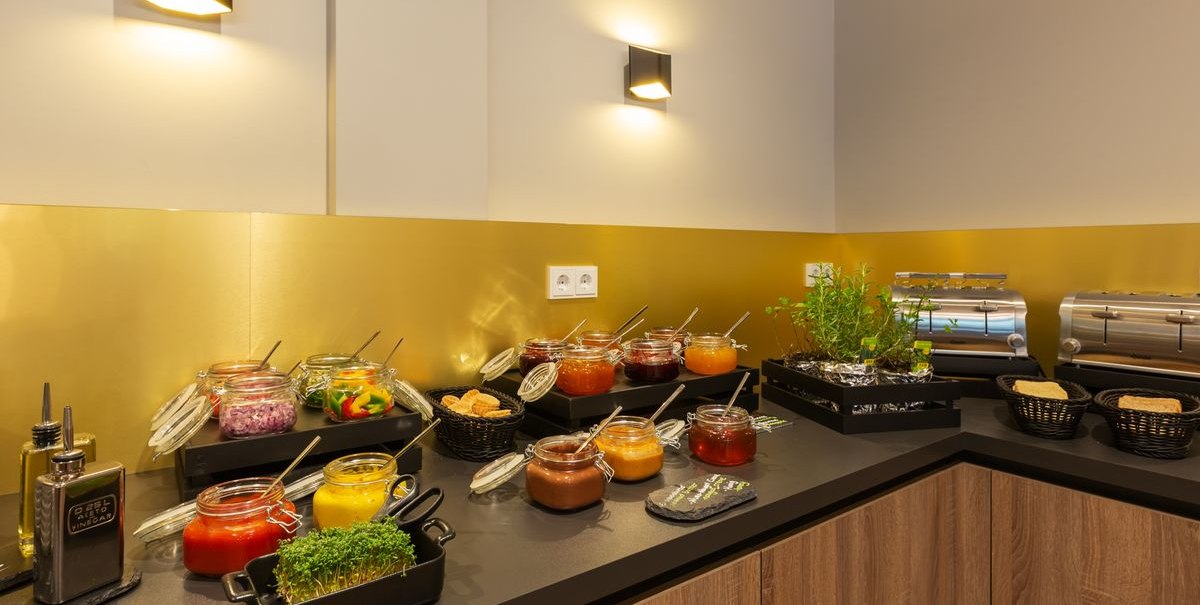 Ein Frühstücksbüffet mit verschiedenen Marmeladen, frischen Kräutern und Toastern auf einer Theke., © Novum Hospitality Ein Frühstücksbüffet mit verschiedenen Marmeladen, frischen Kräutern und Toastern auf einer Theke., © Novum Hospitality