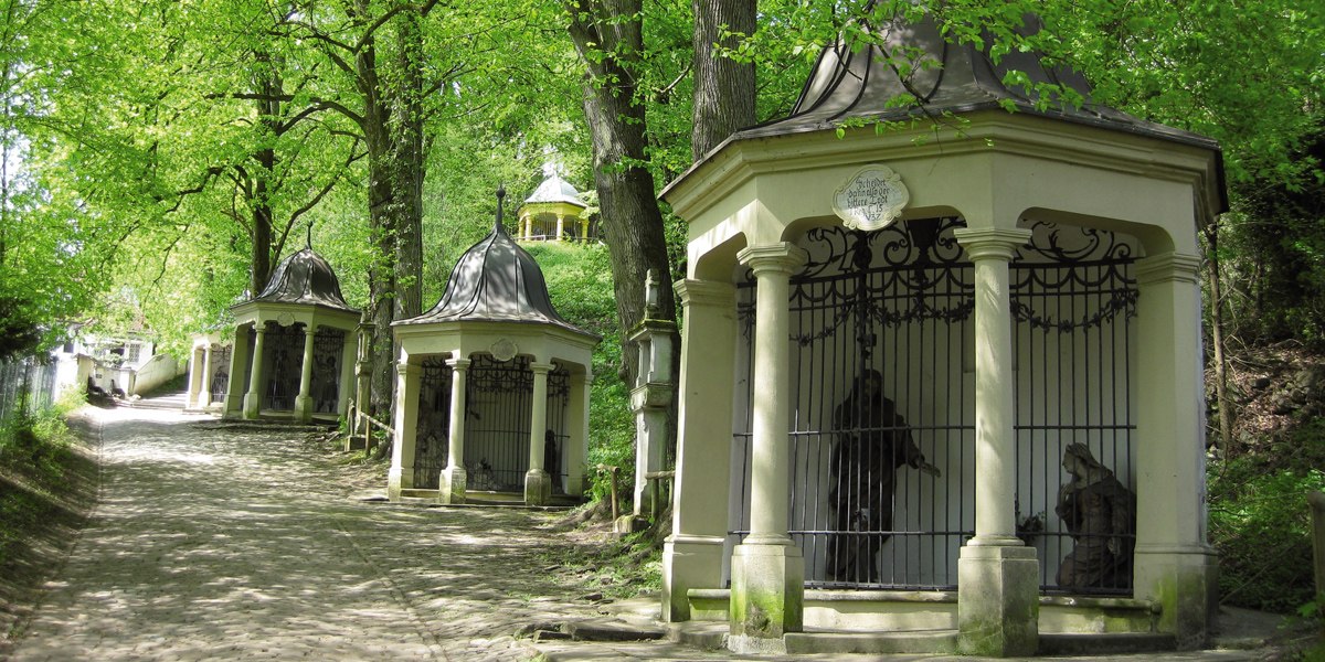 Ein Weg mit mehreren kleinen Kapellen in einem grünen Wald. Die Kapellen haben verzierte Dächer und Gittertüren. Bäume spenden Schatten., © LGS Schwäbisch Gmünd