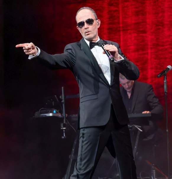 Ein Mann im Anzug und mit Sonnenbrille performt auf einer Bühne vor einem roten Vorhang. Er hält ein Mikrofon und zeigt mit dem Finger nach vorne., © Stadthalle Leonberg Ein Mann im Anzug und mit Sonnenbrille performt auf einer Bühne vor einem roten Vorhang. Er hält ein Mikrofon und zeigt mit dem Finger nach vorne., © Stadthalle Leonberg