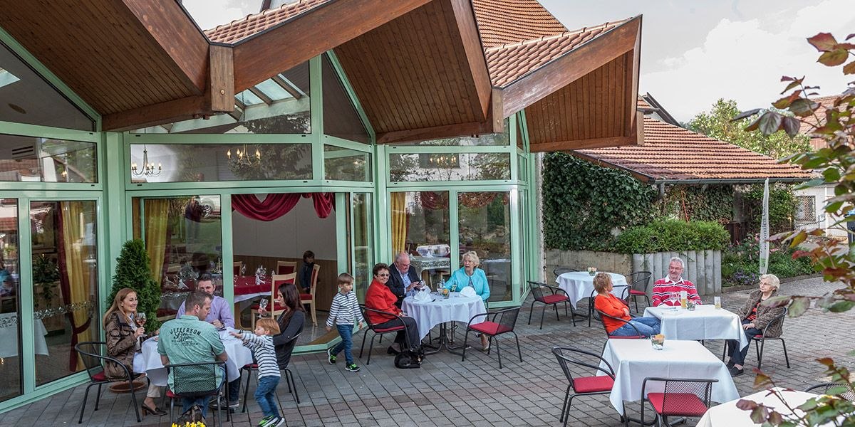 Menschen sitzen an Tischen auf einer Außenterrasse mit großen Fenstern und Holzdach. Kinder spielen, während Erwachsene sich unterhalten., © Hotel Restaurant Lamm