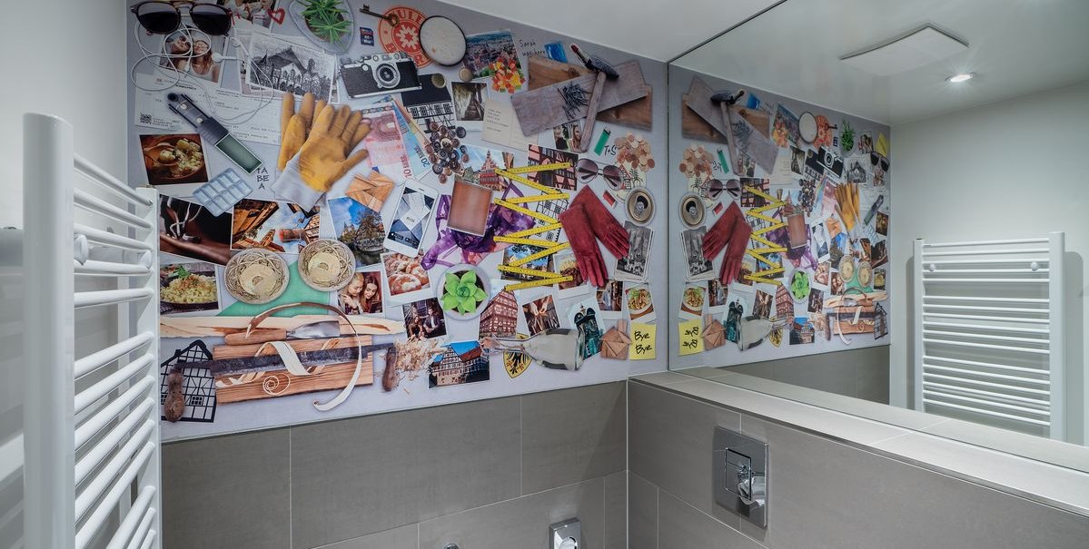 Badezimmer mit bunter Collage aus Fotos und Gegenständen an der Wand, reflektiert im Spiegel. Ein Handtuchhalter ist an der Seite sichtbar., © Novum Hospitality Badezimmer mit bunter Collage aus Fotos und Gegenständen an der Wand, reflektiert im Spiegel. Ein Handtuchhalter ist an der Seite sichtbar., © Novum Hospitality