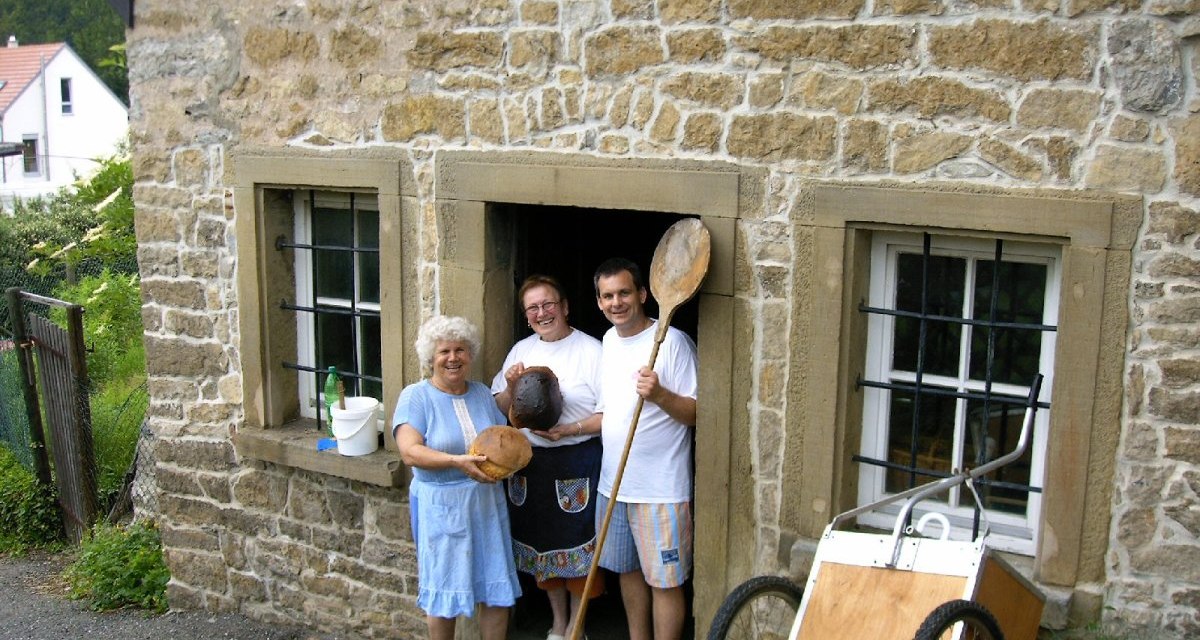 Drei Personen stehen vor einem alten Backhaus aus Stein, halten Brot und Backutensilien. Ein Handwagen lehnt an der Wand., © Land der 1000 Hügel - Kraichgau-Stromberg Drei Personen stehen vor einem alten Backhaus aus Stein, halten Brot und Backutensilien. Ein Handwagen lehnt an der Wand., © Land der 1000 Hügel - Kraichgau-Stromberg