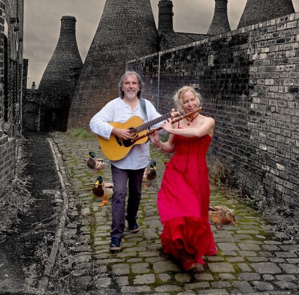 Zwei Musiker, eine Frau in rotem Kleid mit Flöte und ein Mann mit Gitarre, spazieren auf einem Kopfsteinpflasterweg. Enten und alte Schornsteine im Hintergrund., © Mauerwerk GmbH Zwei Musiker, eine Frau in rotem Kleid mit Flöte und ein Mann mit Gitarre, spazieren auf einem Kopfsteinpflasterweg. Enten und alte Schornsteine im Hintergrund., © Mauerwerk GmbH