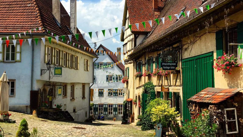 Malerische Altstadtgasse mit Kopfsteinpflaster, Fachwerkhäusern und bunten Wimpeln. Blumen schmücken die Fassaden, sonniges Wetter., © Nürtingen
