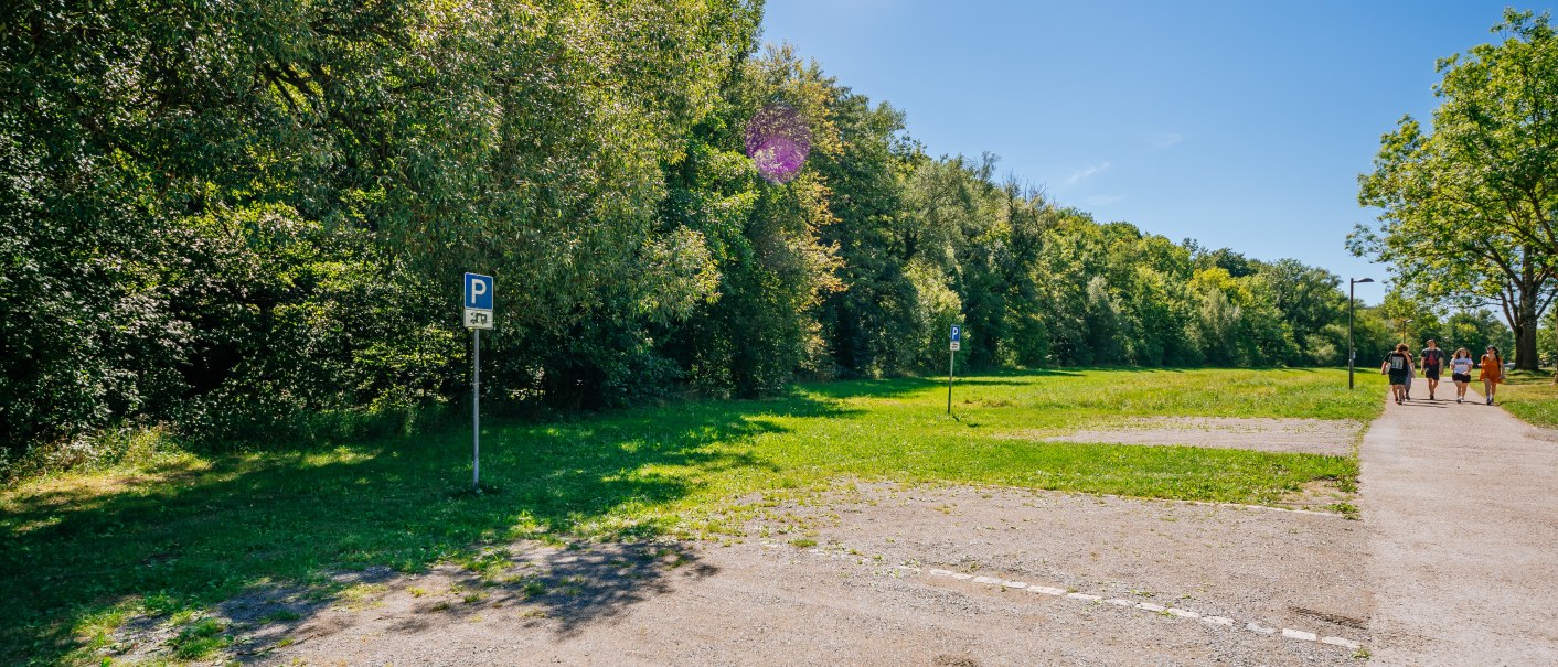 Leerer Wohnmobilstellplatz mit Parkschildern, umgeben von Bäumen. Ein Fußweg führt entlang, auf dem Menschen spazieren. Klarer Himmel., © SMG, Thomas Niedermüller