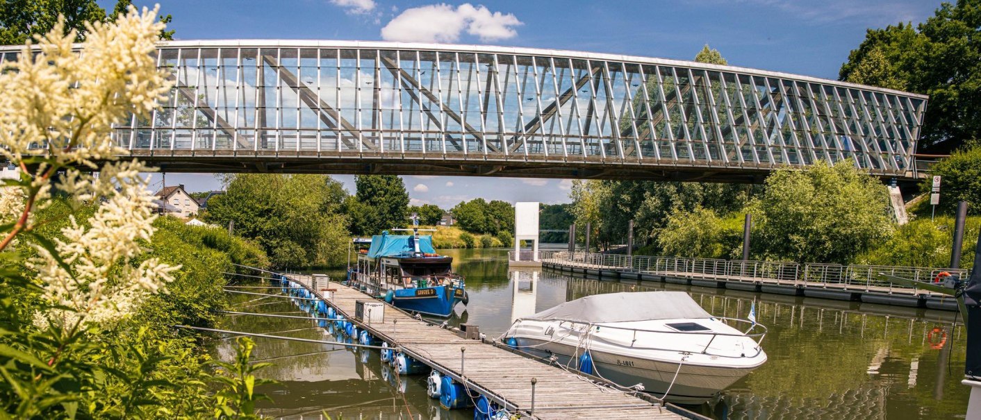 Moderne Glasbrücke über einem Fluss mit Booten und blühenden Pflanzen. Der Himmel ist klar und blau, was eine friedliche Sommeratmosphäre schafft., © Stuttgart-Marketing GmbH, Sarah Schmid