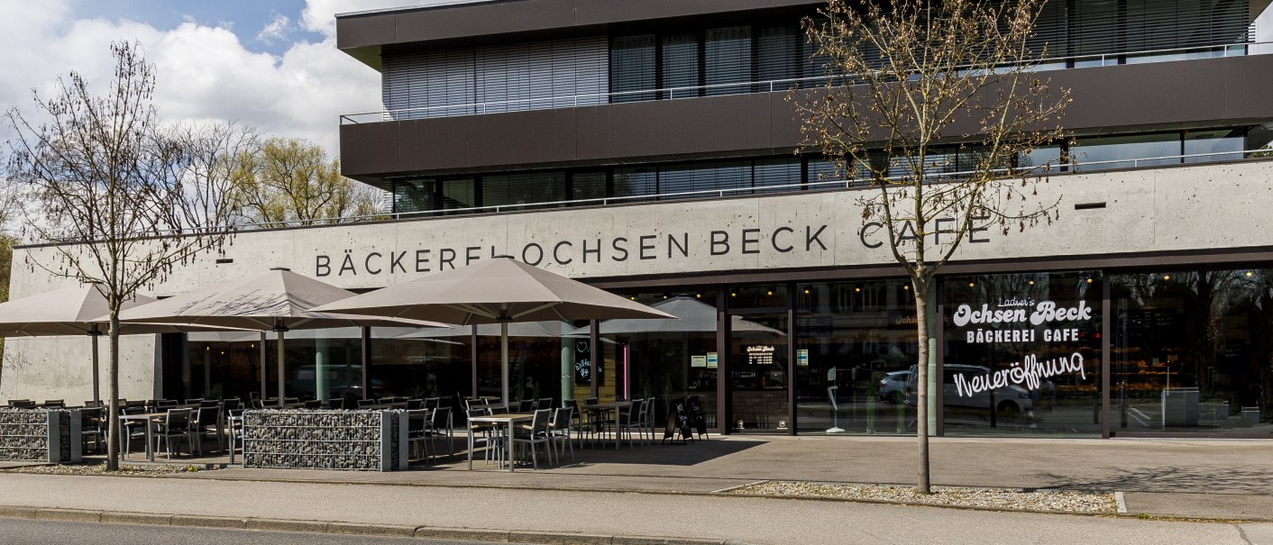 Modernes Café mit Terrasse, Sonnenschirmen und Glasfront. Schriftzug 'Bäckerei Ochsen Beck Café' und Hinweis auf Neueröffnung., © PIET MALL FOTODESIGN Modernes Café mit Terrasse, Sonnenschirmen und Glasfront. Schriftzug 'Bäckerei Ochsen Beck Café' und Hinweis auf Neueröffnung., © PIET MALL FOTODESIGN
