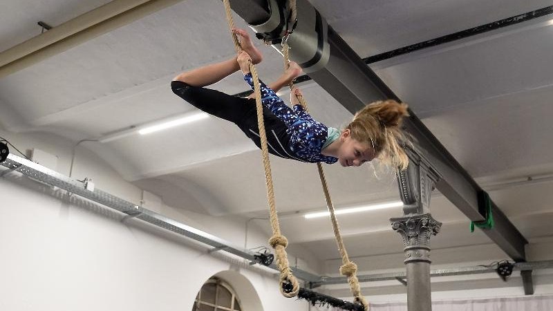 Ein Mädchen in einem blauen Turnanzug macht Luftakrobatik an Seilen in einer Halle mit hohen Decken., © Kunstschule Labyrinth Ein Mädchen in einem blauen Turnanzug macht Luftakrobatik an Seilen in einer Halle mit hohen Decken., © Kunstschule Labyrinth