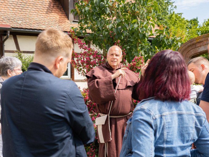 Ein M&ouml;nch in brauner Kutte f&uuml;hrt eine Gruppe von Menschen durch eine malerische Umgebung mit Fachwerkhaus und bl&uuml;henden Pflanzen., &copy; Stadtverwaltung Weil der Stadt