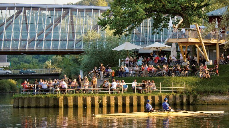 Menschen genießen das Wetter in einem Café am Flussufer, während ein Ruderboot vorbeifährt. Im Hintergrund ist eine moderne Brücke zu sehen., © Michael Fuchs