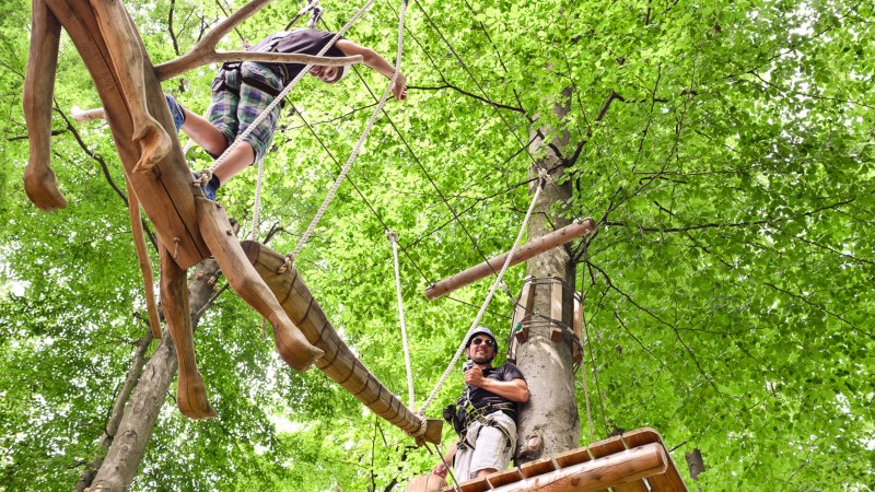 Zwei Personen klettern in einem Hochseilgarten. Eine Person balanciert auf einem Holzsteg, die andere steht auf einer Plattform. Umgeben von gr&uuml;nen Bl&auml;ttern., &copy; Kletterwald Plochingen