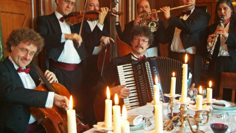 Musiker in festlicher Kleidung spielen Instrumente an einem Tisch mit Kerzenlicht. Die Atmosph&auml;re ist elegant und feierlich.