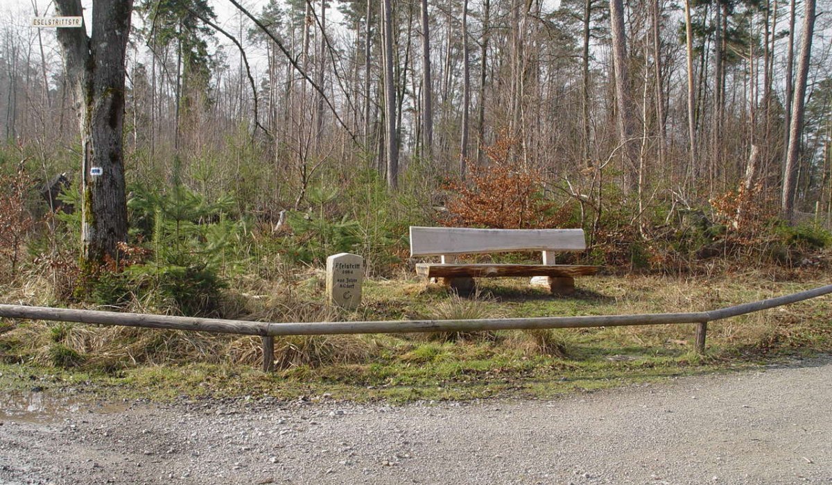 Eine Holzbank steht im Wald neben einem Gedenkstein und einem Wegweiser. Der Boden ist mit Kies bedeckt, und Bäume umgeben die Szene., © Naturpark Schönbuch