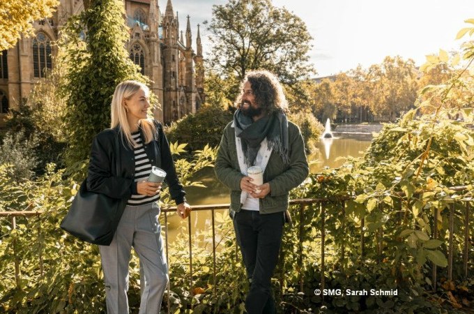 Zwei Personen spazieren lächelnd mit Kaffeebechern vor einer Kirche in Stuttgart. Im Hintergrund ein Teich mit Springbrunnen und herbstlicher Vegetation., © Stuttgart Marketing GmbH