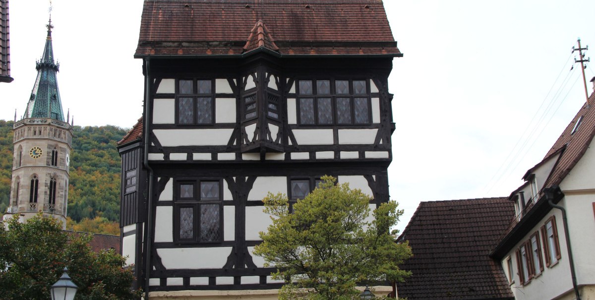 Ein Fachwerkhaus mit markantem Erker, daneben ein Kirchturm. Im Vordergrund ein Baum und eine Straßenlaterne. Ein Fachwerkhaus mit markantem Erker, daneben ein Kirchturm. Im Vordergrund ein Baum und eine Straßenlaterne.