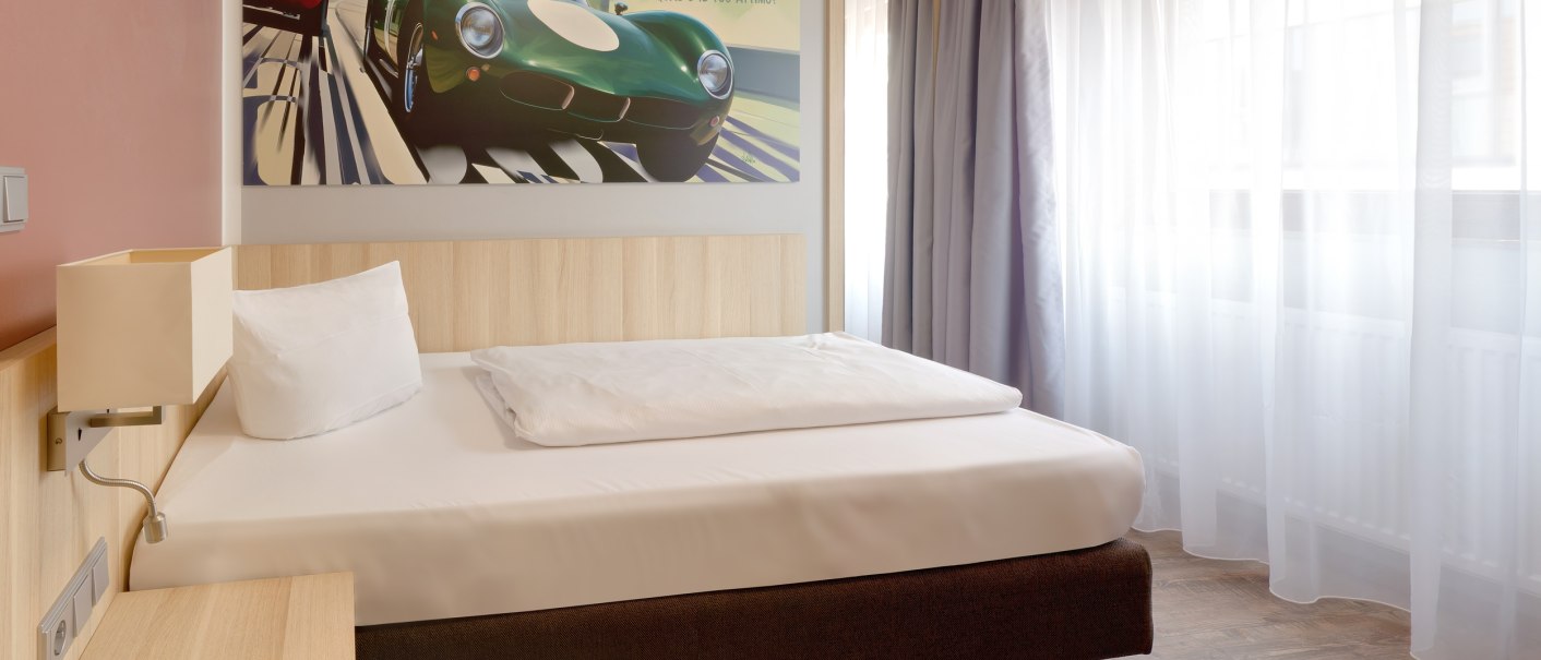 Modernes Einzelzimmer mit Bett, großem Fenster und Vorhängen. An der Wand ein Bild von Autos., © attimo Hotel Stuttgart