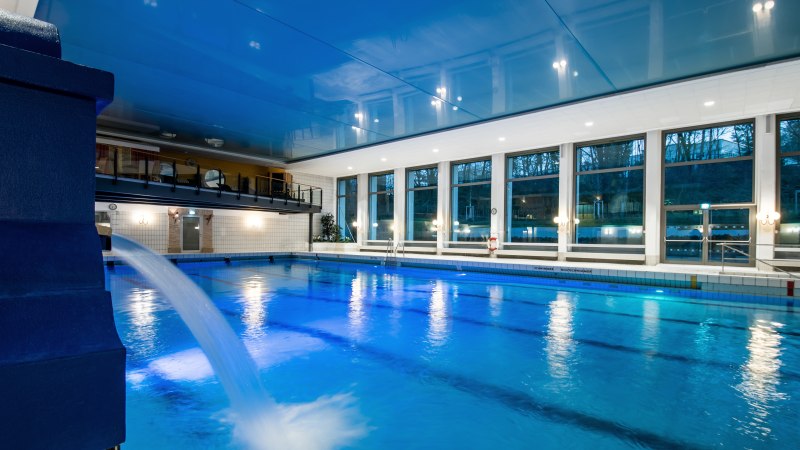 Ein Hallenbad mit blauem Wasser, großen Fenstern und einem Wasserstrahl. Die Decke ist blau, und es gibt eine Galerie im Hintergrund., © Brosch Fotografie / Stadt Wernau