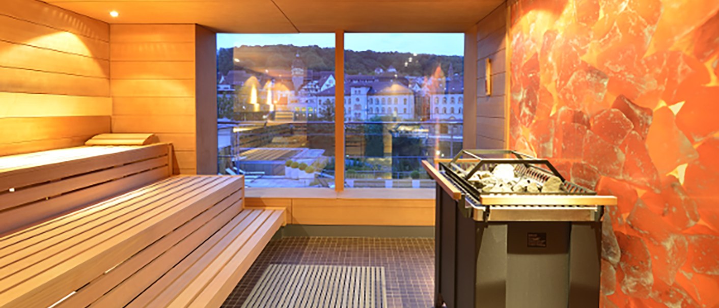 Sauna mit Holzbänken und großem Fenster, das einen Blick auf die Stadt Schwäbisch Hall bietet. Im Vordergrund ein Saunaofen., © Ufuk Arslan