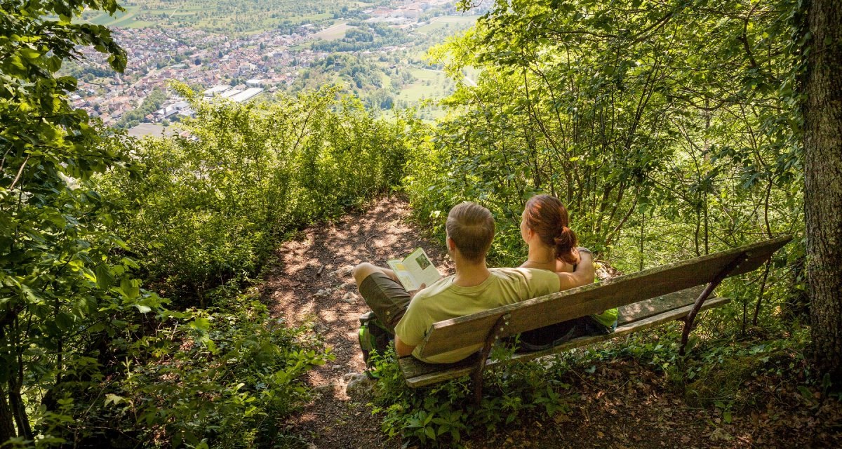 Ein Paar sitzt auf einer Bank im Wald und blickt auf das Lenninger Tal. Die Landschaft ist grün und idyllisch., © hochgehberge Ein Paar sitzt auf einer Bank im Wald und blickt auf das Lenninger Tal. Die Landschaft ist grün und idyllisch., © hochgehberge