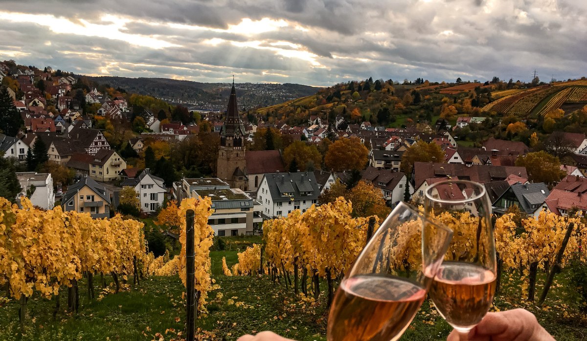 Zwei Personen stoßen mit Weingläsern in einem herbstlichen Weinberg an. Im Hintergrund ist eine Stadt mit Kirche und bunten Bäumen zu sehen., © Stuttgart-Marketing GmbH Zwei Personen stoßen mit Weingläsern in einem herbstlichen Weinberg an. Im Hintergrund ist eine Stadt mit Kirche und bunten Bäumen zu sehen., © Stuttgart-Marketing GmbH