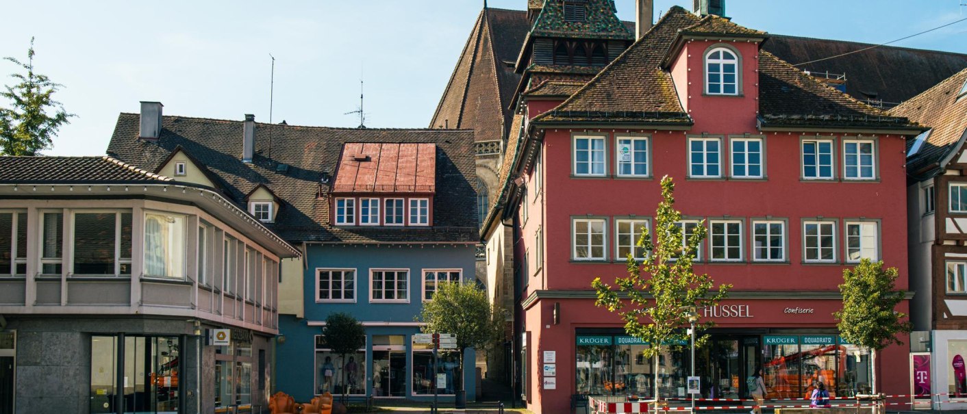 Bunte historische Geb&auml;ude in der Altstadt von Schw&auml;bisch Gm&uuml;nd, darunter ein rotes Haus mit Gesch&auml;ften im Erdgeschoss. Ein Baum steht im Vordergrund., &copy; Stuttgart-Marketing GmbH, Sarah Schmid