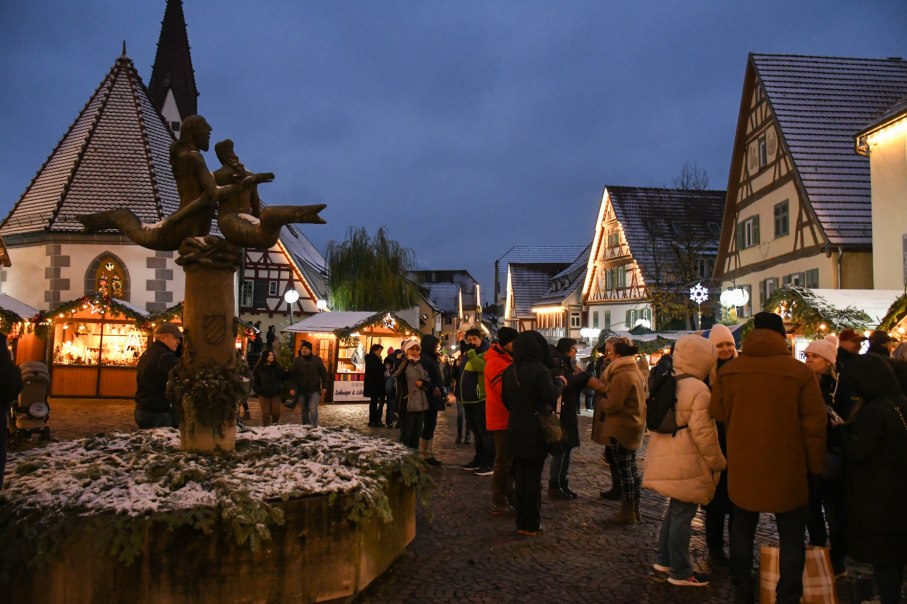 Menschen genießen den Plochinger Weihnachtsmarkt mit beleuchteten Fachwerkhäusern und einer Statue im Vordergrund. Es liegt Schnee auf den Dächern., © Matthias Drißner Menschen genießen den Plochinger Weihnachtsmarkt mit beleuchteten Fachwerkhäusern und einer Statue im Vordergrund. Es liegt Schnee auf den Dächern., © Matthias Drißner