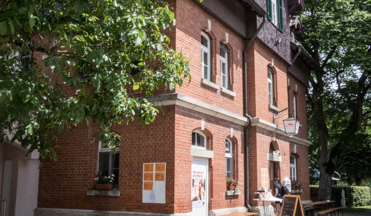 Ein Backsteingeb&auml;ude mit gr&uuml;nen Fensterl&auml;den, umgeben von B&auml;umen. Vor dem Geb&auml;ude stehen Bierb&auml;nke und eine Tafel auf dem Gehweg., &copy; Natur.Nah. Sch&ouml;nbuch & Heckeng&auml;u