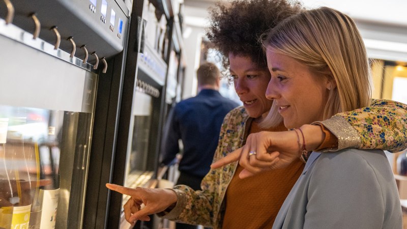 Zwei Frauen stehen vor einem Weinspender und zeigen auf verschiedene Weinflaschen. Im Hintergrund sind weitere Flaschen und Gl&auml;ser zu sehen., &copy; Stuttgart Marketing GmbH, Fotografin Martina Denker