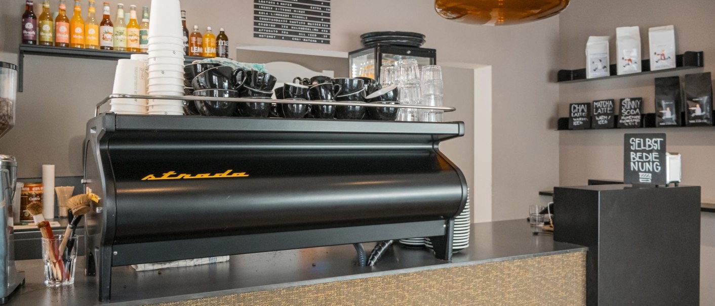 Espressomaschine in einem Café mit schwarzen Tassen, Getränkeflaschen im Hintergrund und einem Schild 'Selbstbedienung'., © SMG, Sarah Schmid Espressomaschine in einem Café mit schwarzen Tassen, Getränkeflaschen im Hintergrund und einem Schild 'Selbstbedienung'., © SMG, Sarah Schmid