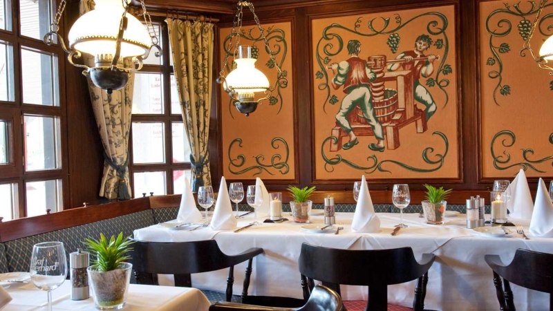 Elegantes Restaurant mit gedecktem Tisch, wei&szlig;en Servietten, Wandmalerei von Weinbauern und Kronleuchtern. Gem&uuml;tliche Atmosph&auml;re mit Holzelementen., &copy; Biosph&auml;renhotel Graf Eberhard