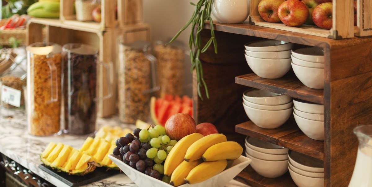 Ein Frühstücksbuffet mit frischem Obst wie Bananen, Trauben und Äpfeln, sowie Müsli in Behältern und gestapelten Schalen auf einem Marmortisch., © Novum Hospitality
