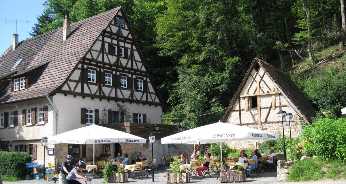 Ein Fachwerkhaus mit Biergarten, umgeben von Bäumen. Menschen sitzen an Tischen, während zwei Radfahrer im Vordergrund stehen., © Natur.Nah. Schönbuch & Heckengäu Ein Fachwerkhaus mit Biergarten, umgeben von Bäumen. Menschen sitzen an Tischen, während zwei Radfahrer im Vordergrund stehen., © Natur.Nah. Schönbuch & Heckengäu