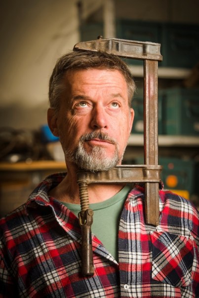 Ein Mann mit Bart und kariertem Hemd trägt eine große Schraubzwinge um den Hals. Er blickt nach oben. Im Hintergrund sind Regale zu sehen., © Kulturhaus Schwanen Ein Mann mit Bart und kariertem Hemd trägt eine große Schraubzwinge um den Hals. Er blickt nach oben. Im Hintergrund sind Regale zu sehen., © Kulturhaus Schwanen