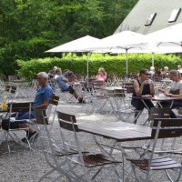 Menschen sitzen im Außenbereich eines Cafés mit weißen Sonnenschirmen, umgeben von Bäumen und grüner Vegetation., © Gabriel Holom