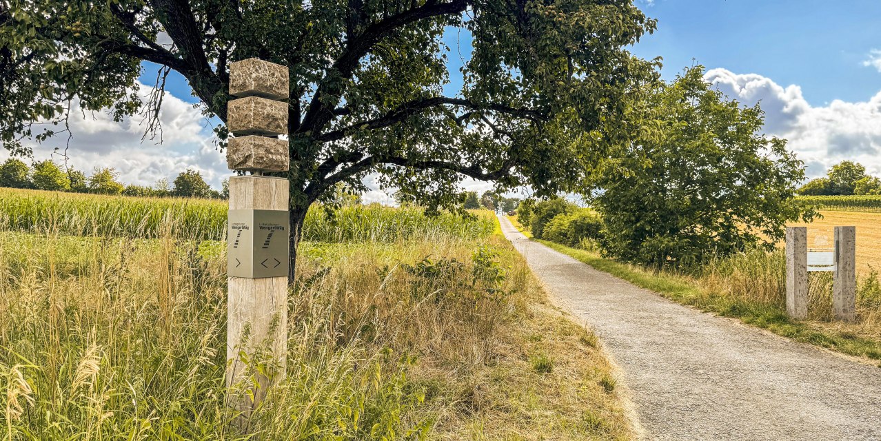 Ein Wegweiser aus Stein und Holz steht neben einem Feldweg, umgeben von B&auml;umen und Feldern unter einem blauen Himmel mit Wolken., &copy; SMG, Ellen Haeffner
