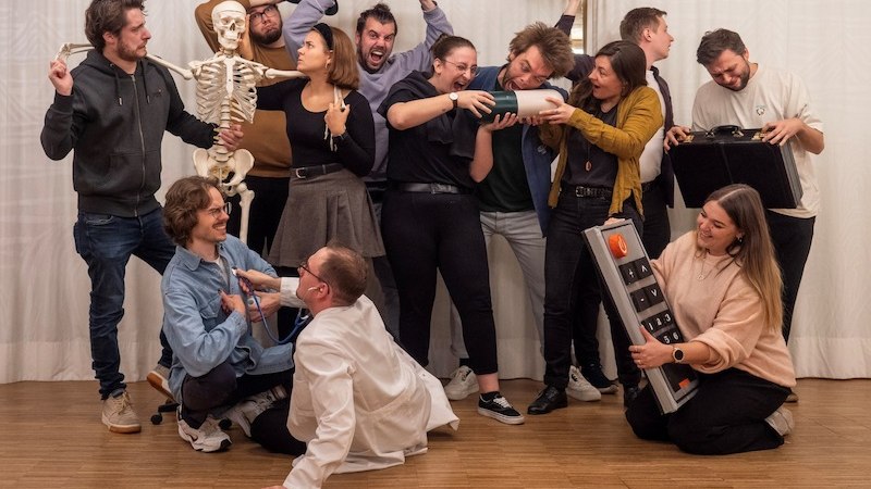 Eine Gruppe von Menschen posiert humorvoll mit einem Skelett und übergroßen Requisiten in einem Raum mit Holzboden., © Kulturhaus Schwanen Eine Gruppe von Menschen posiert humorvoll mit einem Skelett und übergroßen Requisiten in einem Raum mit Holzboden., © Kulturhaus Schwanen