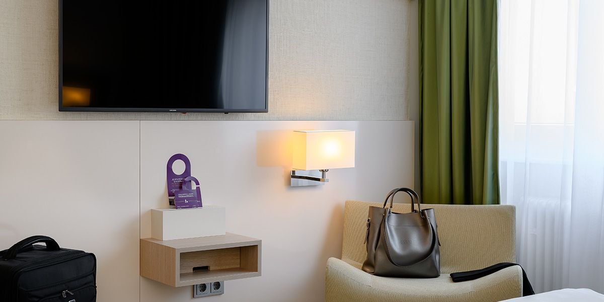 Ein modernes Hotelzimmer mit einem großen Fernseher, einer Wandlampe, einem Sessel mit Tasche und grünen Vorhängen., © Mercure Bristol Stuttgart Sindelfingen