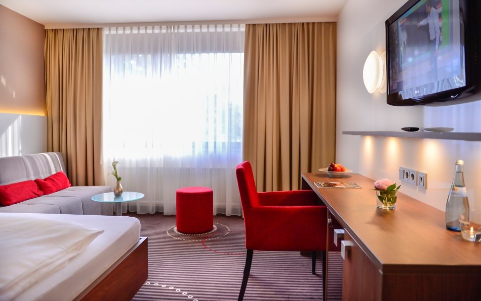 Modernes Hotelzimmer mit Doppelbett, Sofa, Schreibtisch, TV und großen Fenstern. Rote Akzente und dezente Beleuchtung schaffen eine gemütliche Atmosphäre., © Hotel Fuchsen Modernes Hotelzimmer mit Doppelbett, Sofa, Schreibtisch, TV und großen Fenstern. Rote Akzente und dezente Beleuchtung schaffen eine gemütliche Atmosphäre., © Hotel Fuchsen