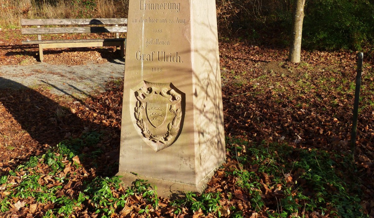 Ein Gedenkstein im Wald mit Inschrift und Wappen, umgeben von Laub und Pflanzen. Im Hintergrund steht eine Bank., © Natur.Nah. Schönbuch & Heckengäu