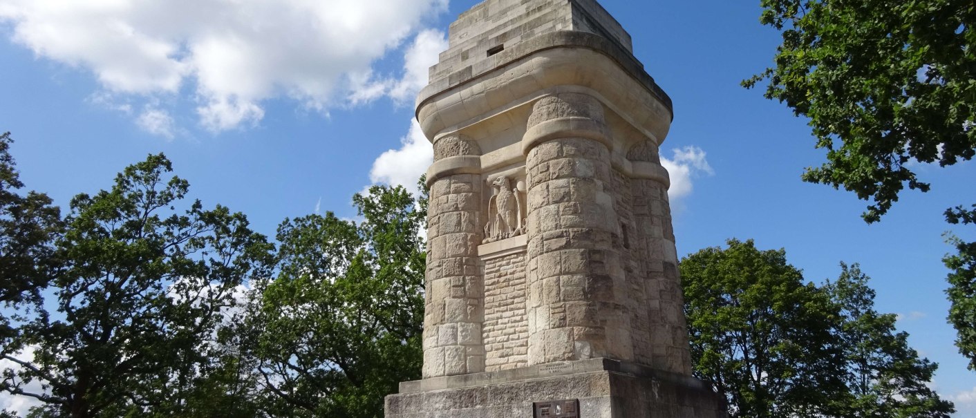 Ein massiver Bismarckturm aus Stein steht auf einem Platz, umgeben von Bäumen und unter einem blauen Himmel mit wenigen Wolken., © SMG